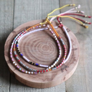 May include: Three beaded bracelets with adjustable cords rest on a wooden surface. The bracelets feature small, colorful beads in various patterns, including red, white, black, and gold. The cords are in shades of yellow, pink, and red.