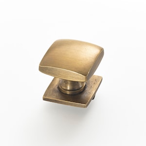 May include: A square cabinet knob with a brushed brass finish. The knob has a raised edge and a slightly rounded top.