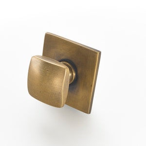 May include: A square-shaped, brushed brass cabinet knob with a square-shaped, brushed brass base.