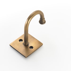 May include: A brass wall-mounted hook with a curved arm and a square base. The hook is designed for hanging items.