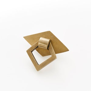 May include: A brass cabinet pull with a square opening and a diamond-shaped base.