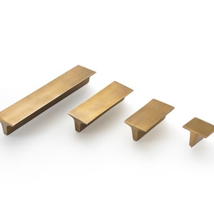 May include: Four brushed brass cabinet pulls in various sizes. The pulls have a rectangular shape with a T-shaped base.