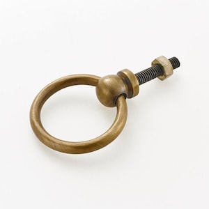 Può includere: Una maniglia ad anello di trazione in ottone con un pomo rotondo e una vite. La maniglia è di colore oro chiaro.