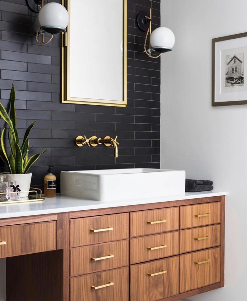 May include: A bathroom vanity with a white countertop and a white porcelain sink. The vanity has six drawers with gold handles and is made of wood with a dark brown finish. The vanity is mounted to the wall and has a black tile backsplash with gold accents.