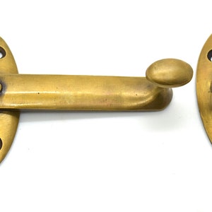 May include: A brass window sash lock set against a white background. The set includes two oval-shaped pieces with screw holes and a pivoting arm with a rounded handle. The metal has a warm, aged finish.