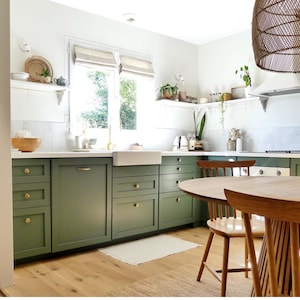 May include: A kitchen with green cabinets, white countertops, and a white farmhouse sink. The kitchen has a wood floor and a round wooden table with two chairs. There are plants on shelves and a wicker pendant light hanging over the table.