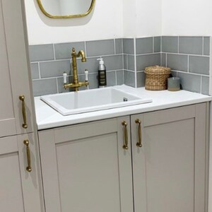 May include: A bathroom vanity with a white sink, brass faucet, and gray cabinets. A gold-framed mirror hangs above the sink, with gray tile backsplash. A woven basket and candle sit on the countertop.