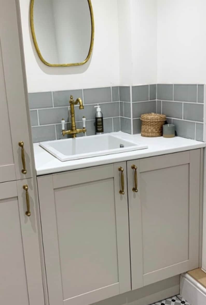 May include: A bathroom vanity with a white sink, brass faucet, and gray cabinets. A gold-framed mirror hangs above the sink, with gray tile backsplash. A woven basket and candle sit on the countertop.