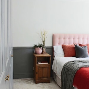 May include: Bedroom interior with a pink button-tufted headboard, white bedding, and rust-colored pillows. A wooden nightstand holds a pink pot with plants and a decorative vase. A gray blanket and red bedspread complete the cozy scene.