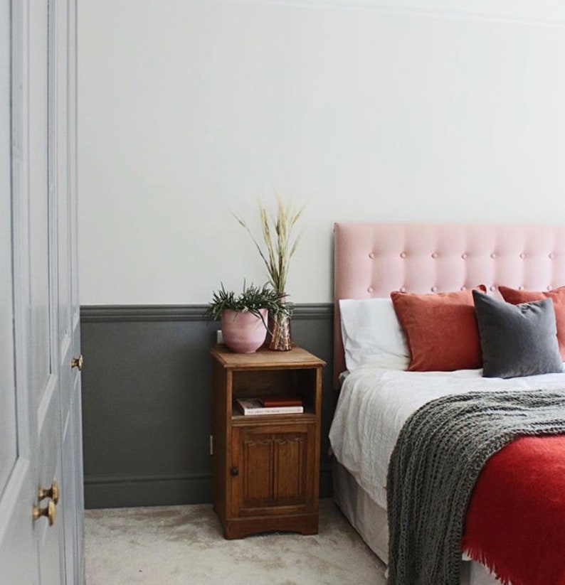May include: Bedroom interior with a pink button-tufted headboard, white bedding, and rust-colored pillows. A wooden nightstand holds a pink pot with plants and a decorative vase. A gray blanket and red bedspread complete the cozy scene.