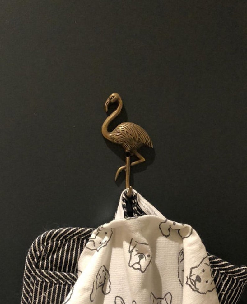 May include: A bronze flamingo hook mounted on a dark wall, holding a white towel with a black dog print and a black and white striped towel. The hook is a decorative accent.