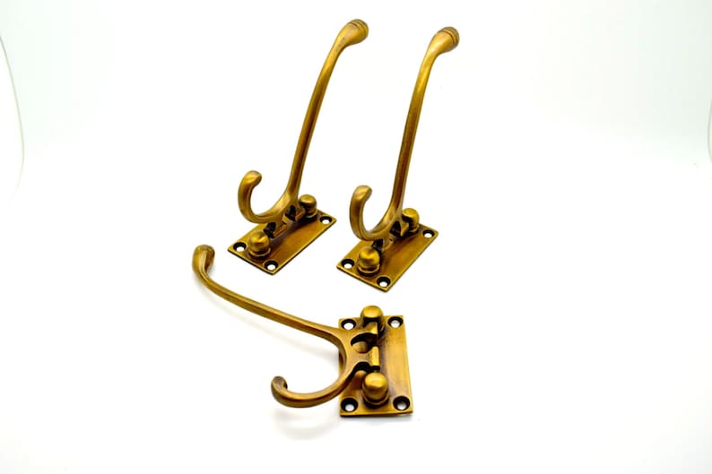 May include: Three antique brass coat hooks. Each hook has a curved design and is mounted on a rectangular base with screw holes. The hooks are a warm, golden color, suggesting an aged finish. Ideal for entryways or closets.