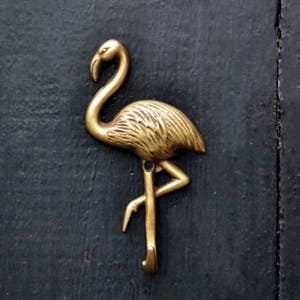 May include: A brass flamingo-shaped wall hook. The hook is a golden color and is mounted on a dark blue wooden surface. The flamingo has a long, curved neck and a detailed body.