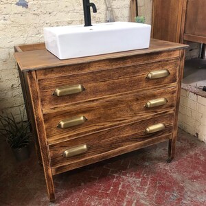 May include: A wooden bathroom vanity with four drawers and brass pulls. The vanity has a white porcelain sink and a black faucet.