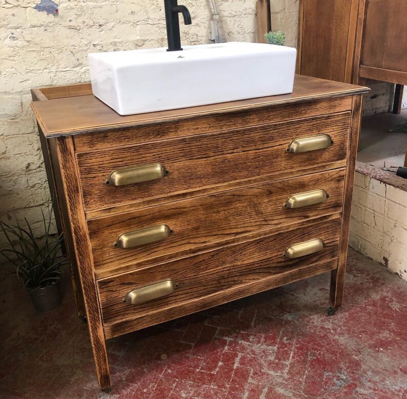 May include: A wooden bathroom vanity with four drawers and brass pulls. The vanity has a white porcelain sink and a black faucet.