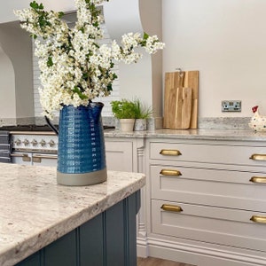 May include: A blue and gray ceramic pitcher filled with white flowers sits on a white countertop in a kitchen. The countertop is made of granite and has a speckled pattern. The pitcher is sitting in front of a set of white cabinets with gold drawer pulls.