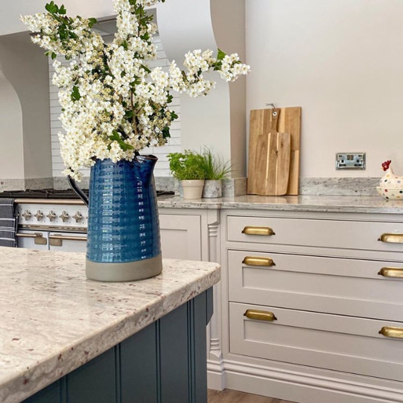 May include: A blue and gray ceramic pitcher filled with white flowers sits on a white countertop in a kitchen. The countertop is made of granite and has a speckled pattern. The pitcher is sitting in front of a set of white cabinets with gold drawer pulls.