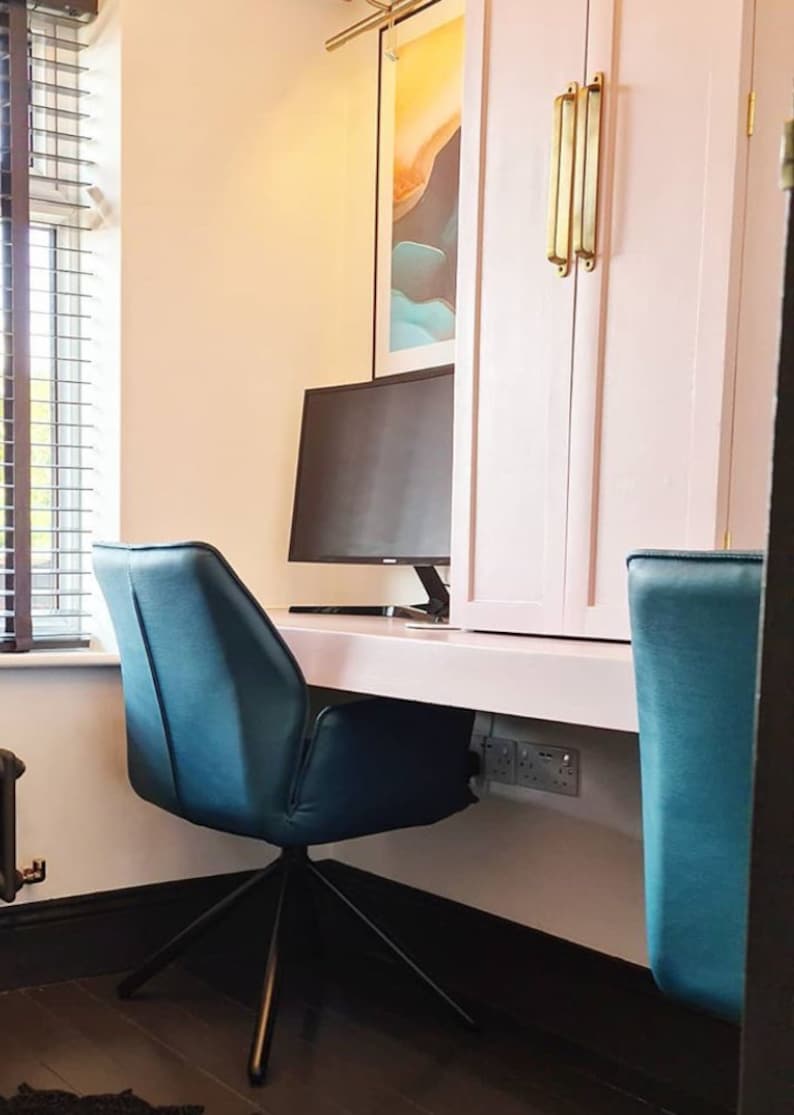 May include: A white desk with a black computer monitor and two teal blue office chairs. The desk is mounted to the wall and has a white cabinet above it. The cabinet has two doors with gold handles.