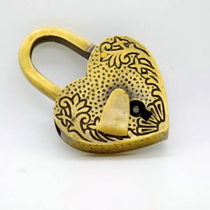 May include: A heart-shaped brass padlock with an ornate floral design. The lock has a curved handle and a keyhole. The surface is textured with small dots. The overall color is a warm, antique gold.