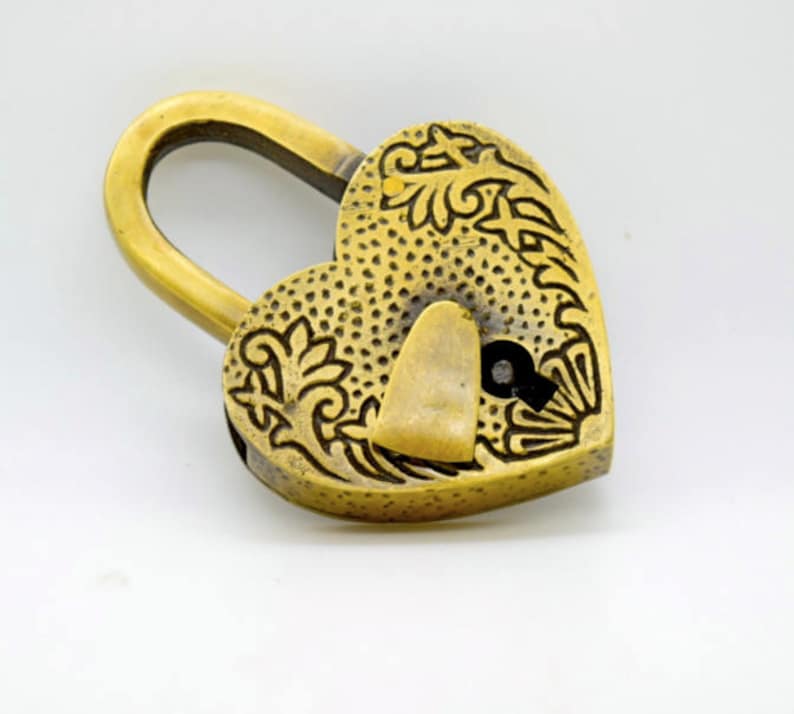 May include: A heart-shaped brass padlock with an ornate floral design. The lock has a curved handle and a keyhole. The surface is textured with small dots. The overall color is a warm, antique gold.