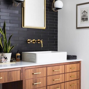 May include: A bathroom vanity with a white countertop and a white porcelain sink. The vanity has six drawers with gold handles and is made of wood with a dark brown finish. The vanity is mounted to the wall and has a black tile backsplash with gold accents.