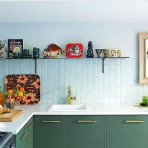 May include: A kitchen with green cabinets, a white countertop, and a light blue backsplash. A glass shelf displays various decorative items. A framed abstract artwork hangs on the wall, and a cutting board with bread sits on the counter.