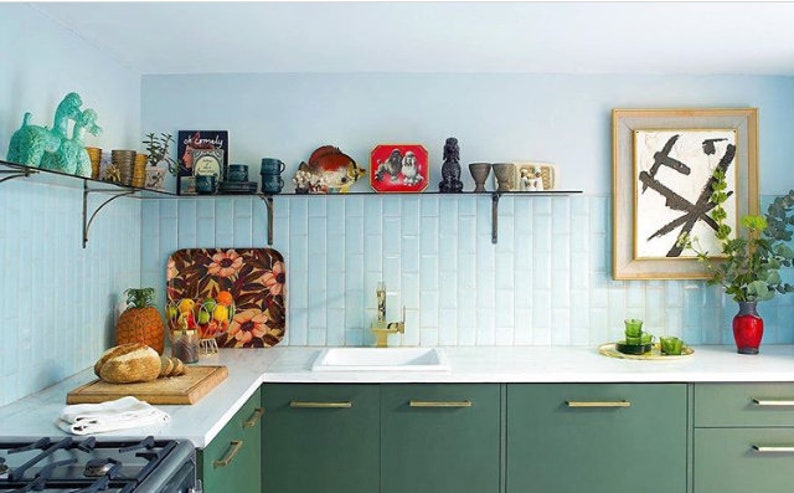 May include: A kitchen with green cabinets, a white countertop, and a light blue backsplash. A glass shelf displays various decorative items. A framed abstract artwork hangs on the wall, and a cutting board with bread sits on the counter.