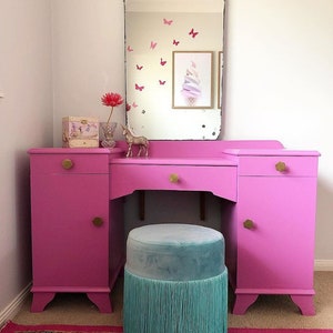 May include: A pink vanity set with a large mirror, two drawers, and a round, turquoise velvet stool with a fringe trim. The vanity has gold knobs and is set against a white wall.