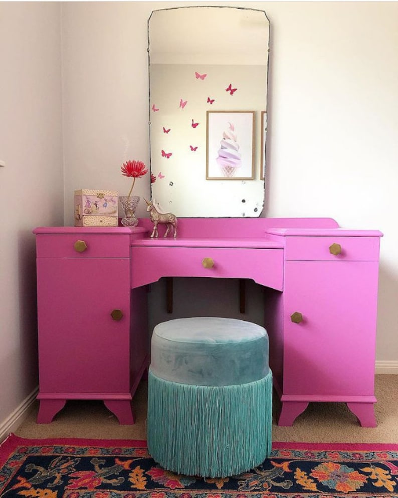 May include: A pink vanity set with a large mirror, two drawers, and a round, turquoise velvet stool with a fringe trim. The vanity has gold knobs and is set against a white wall.