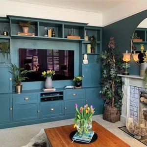 May include: A teal-colored built-in entertainment center with a large TV, cabinets, and shelves. The room features a fireplace, a round mirror, and a wooden coffee table with a vase of tulips. Decorative items include plants, candles, and books.