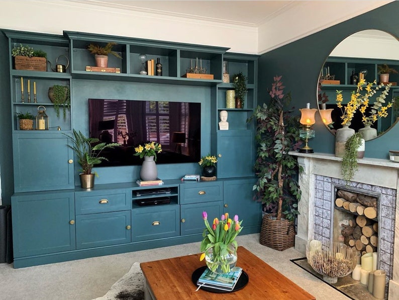 May include: A teal-colored built-in entertainment center with a large TV, cabinets, and shelves. The room features a fireplace, a round mirror, and a wooden coffee table with a vase of tulips. Decorative items include plants, candles, and books.