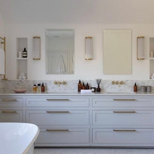 May include: A modern bathroom with a white bathtub and a double vanity. The vanity has light gray cabinets with gold hardware. Above the vanity are mirrors, sconces, and gold towel racks. The countertop is marble, and the walls are white.
