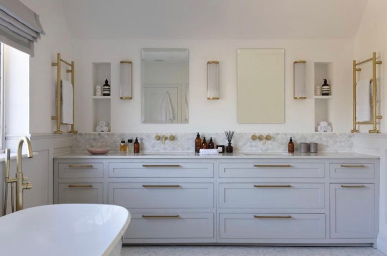 May include: A modern bathroom with a white bathtub and a double vanity. The vanity has light gray cabinets with gold hardware. Above the vanity are mirrors, sconces, and gold towel racks. The countertop is marble, and the walls are white.