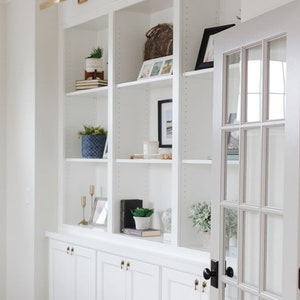May include: White built-in bookshelves with a glass door, featuring decorative items such as plants, books, and framed photos. The shelves are accented with gold light fixtures and black door hardware.
