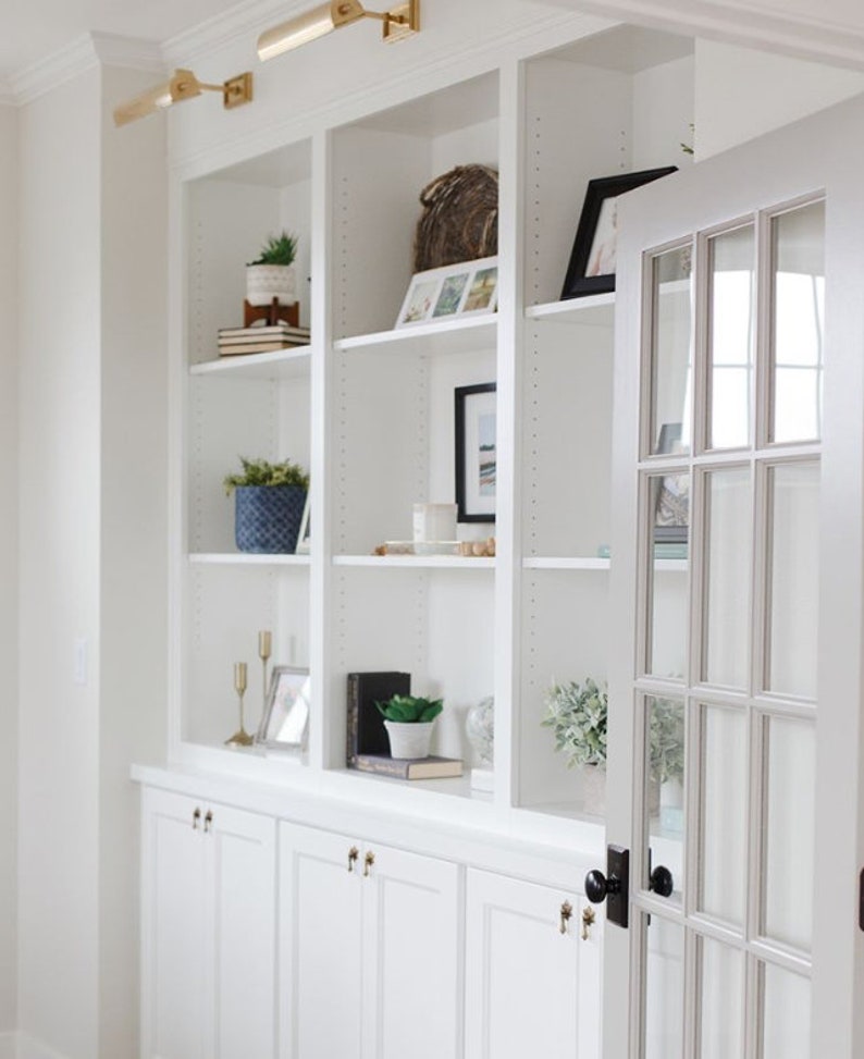 May include: White built-in bookshelves with a glass door, featuring decorative items such as plants, books, and framed photos. The shelves are accented with gold light fixtures and black door hardware.