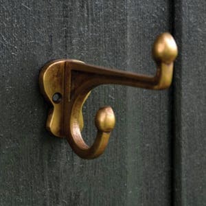 May include: A brass-colored wall hook with a curved design. The hook is mounted on a dark green wooden surface. The hook has a rounded top and a curved lower hook.