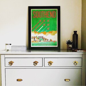 May include: A vintage travel poster with a black frame, featuring a green background with orange text that reads "SOUTHEND". The poster depicts a cityscape with a pier and several red airplanes flying in formation.