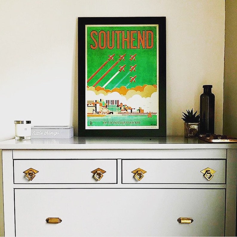 May include: A vintage travel poster with a black frame, featuring a green background with orange text that reads "SOUTHEND". The poster depicts a cityscape with a pier and several red airplanes flying in formation.