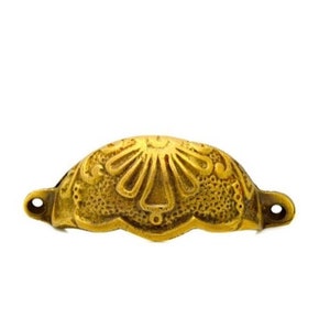 May include: A gold-toned metal drawer pull with an ornate, floral design. The pull is shaped like a half-circle and has a raised, textured surface.