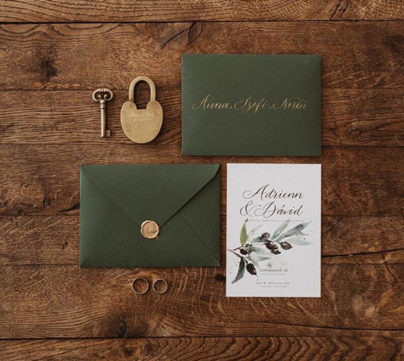 May include: Wedding invitation suite featuring a gold key, lock, and rings, with green envelopes and a card with the names Adrienn & D&aacute;vid. The invitation suite is arranged on a wooden surface.