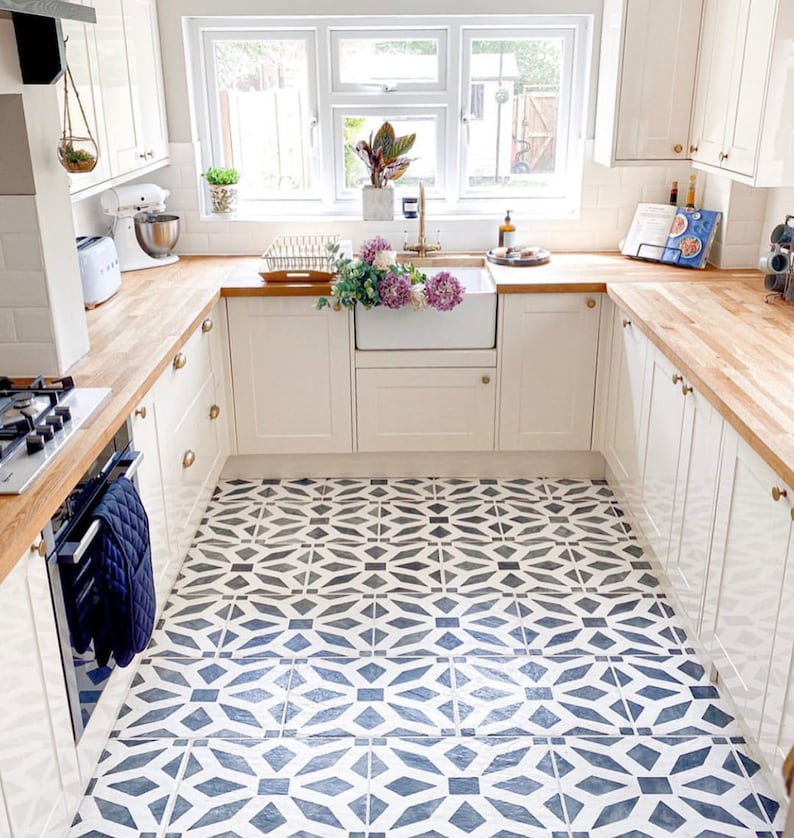May include: A white kitchen with wood countertops and white cabinets. The floor is tiled with a black and white geometric pattern. There is a window with a view of a garden.