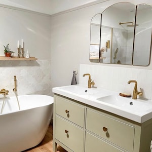 May include: A modern bathroom with a white freestanding bathtub, a double sink vanity with gold fixtures, and a large mirror. The vanity is a light green color with gold knobs. The walls are white with decorative tile.