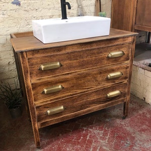 May include: A wooden bathroom vanity with four drawers and brass pulls. The vanity has a white porcelain sink and a black faucet.