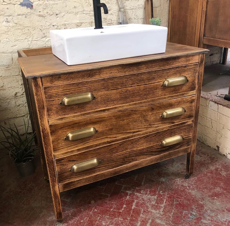May include: A wooden bathroom vanity with four drawers and brass pulls. The vanity has a white porcelain sink and a black faucet.
