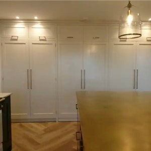 May include: A white kitchen with a large set of white cabinets with long, horizontal handles. The cabinets are closed and have a smooth, flat surface. The cabinets are in a row and are all the same size. The floor is made of light wood and has a herringbone pattern. A large, gold countertop is in the foreground.