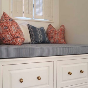 May include: A white window seat with a blue and white striped cushion. The seat has two white drawers with gold knobs. There are three pillows on the seat, two with red and orange patterns and one with a blue and white pattern.