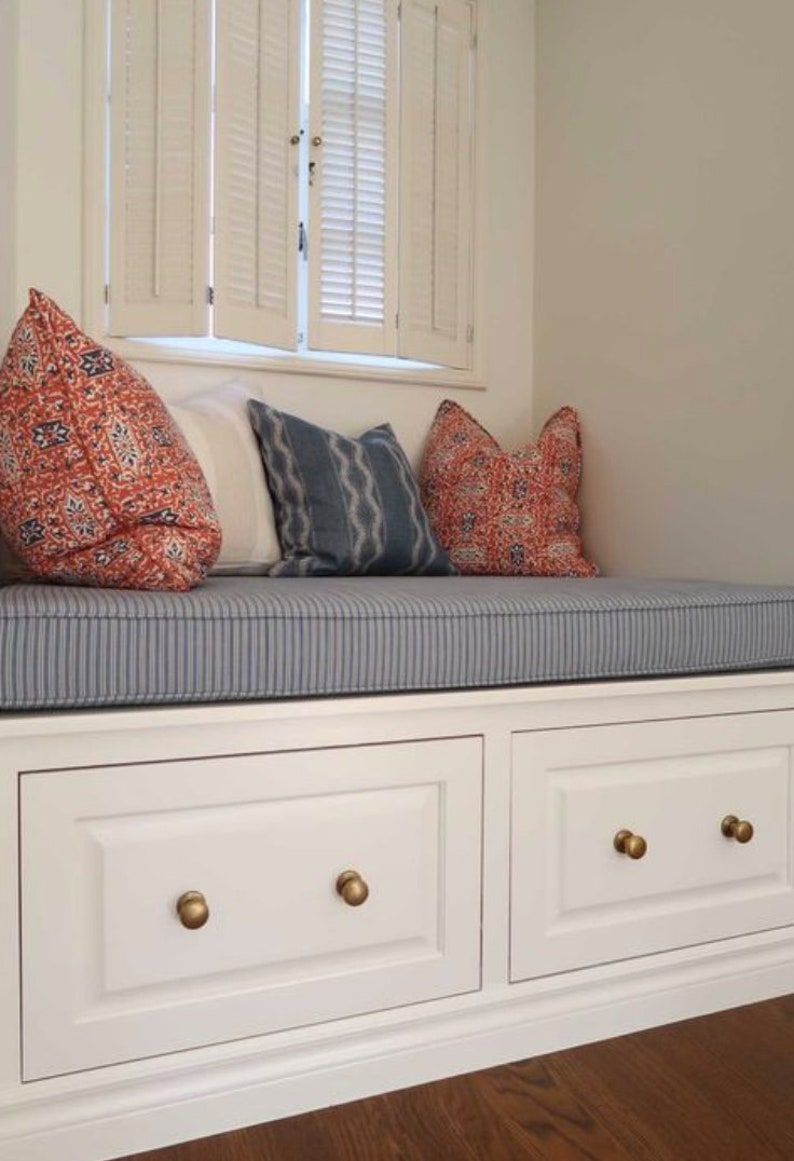 May include: A white window seat with a blue and white striped cushion. The seat has two white drawers with gold knobs. There are three pillows on the seat, two with red and orange patterns and one with a blue and white pattern.