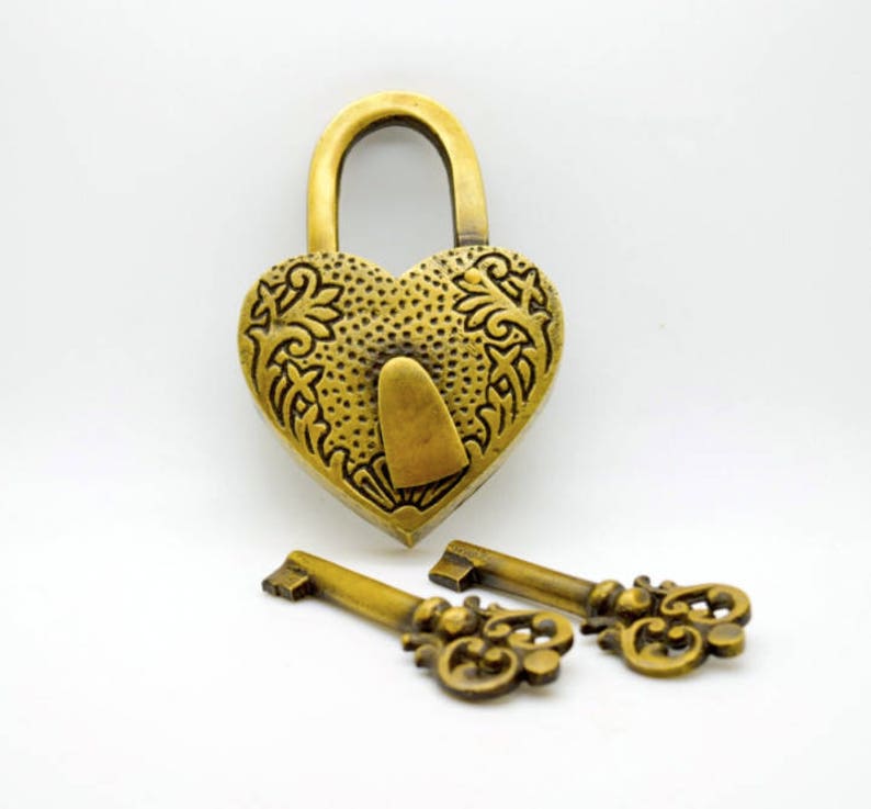 May include: A heart-shaped, antique brass padlock with ornate floral detailing and a matching brass key. The lock is closed, and two keys are placed below it. The lock and keys are on a white background.