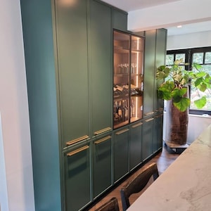 May include: A built-in, dark green cabinet with gold-toned handles. A glass-fronted wine cabinet is in the center, illuminated with interior lighting. The cabinets are part of a kitchen or dining area.