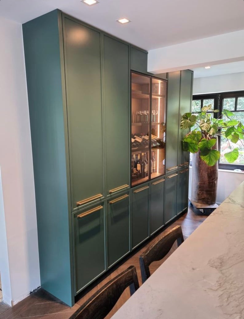May include: A built-in, dark green cabinet with gold-toned handles. A glass-fronted wine cabinet is in the center, illuminated with interior lighting. The cabinets are part of a kitchen or dining area.